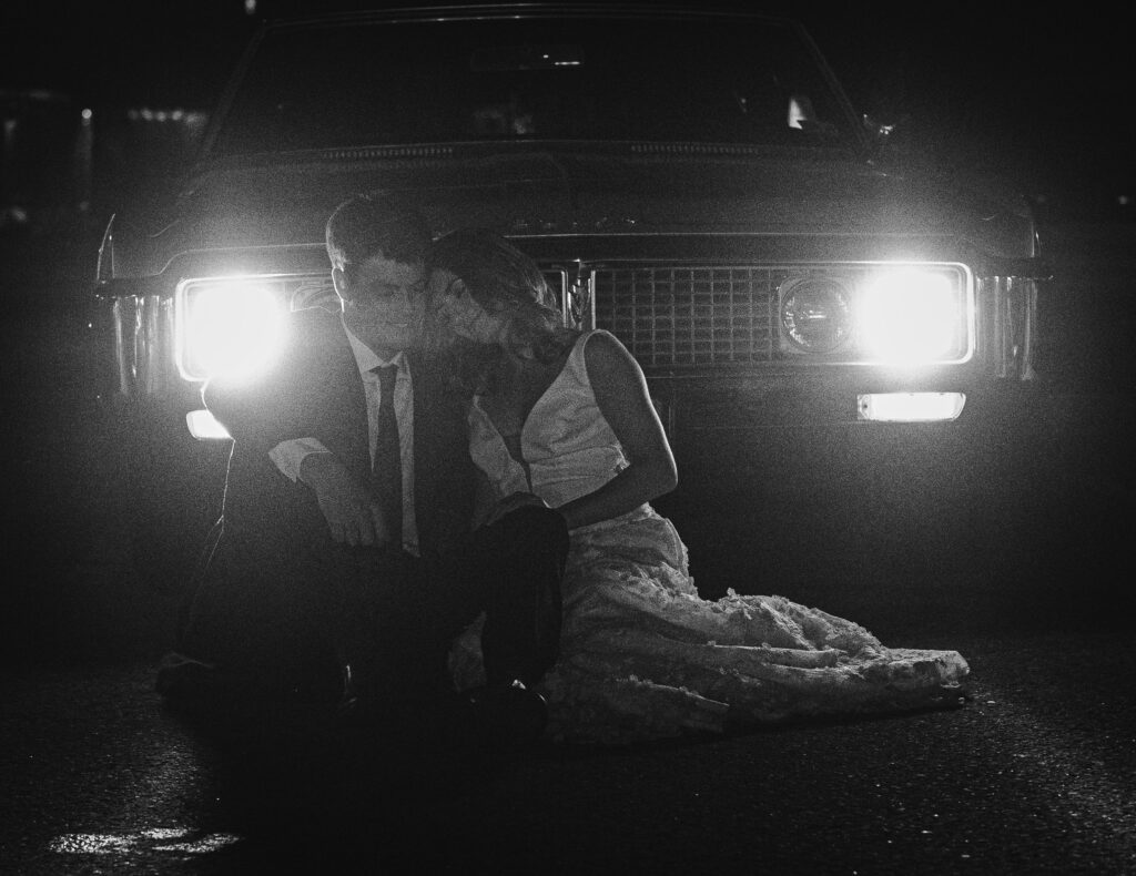 bride and groom sitting in front of the cars headlights