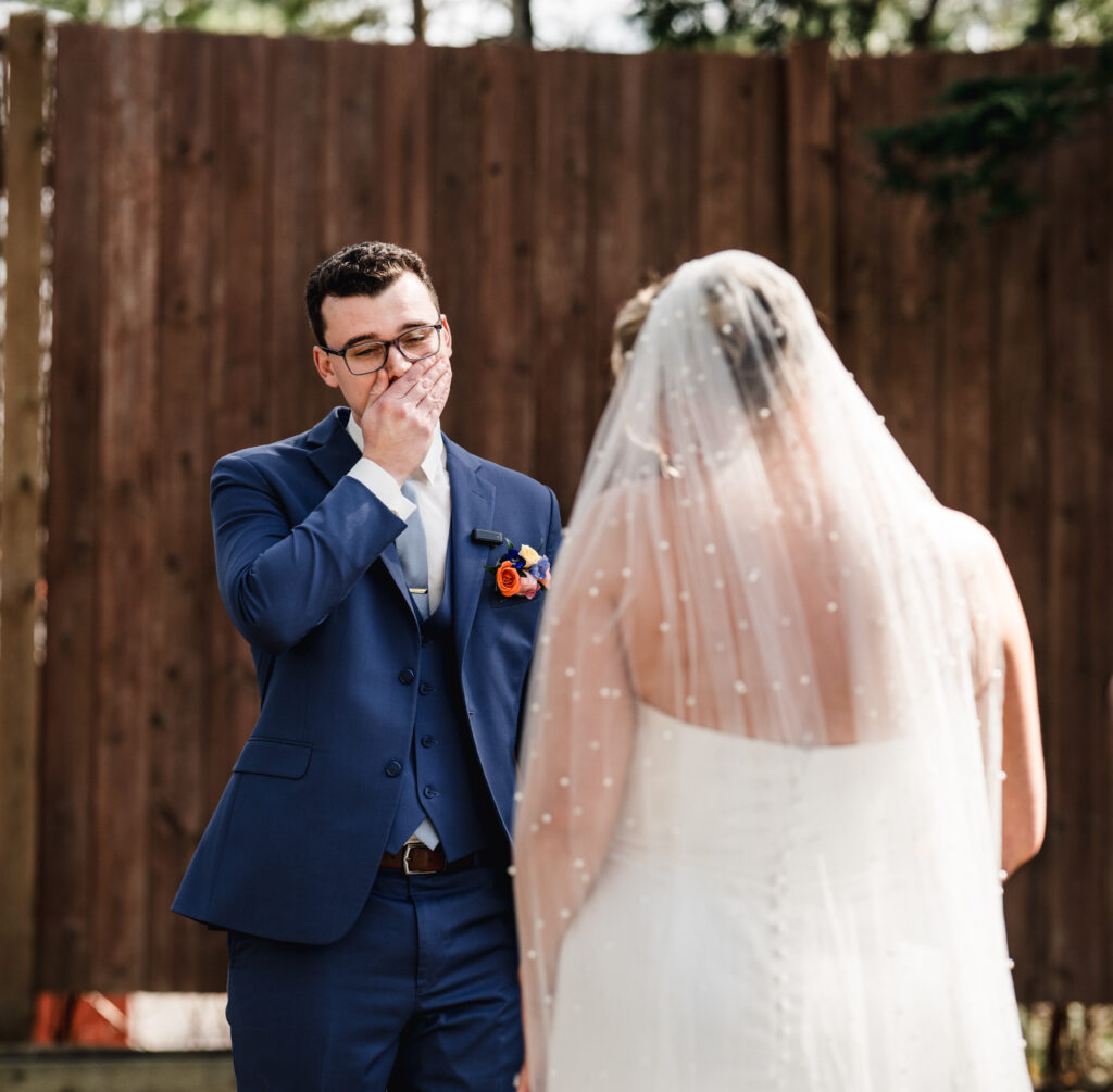An emotional groom, dressed in a navy suit with vibrant florals pinned to his lapel, covers his mouth in awe as he sees his bride for the first time. The bride, wearing a pearl-dotted veil and strapless gown, stands facing him, capturing a powerful first look moment filled with raw, heartfelt emotion
