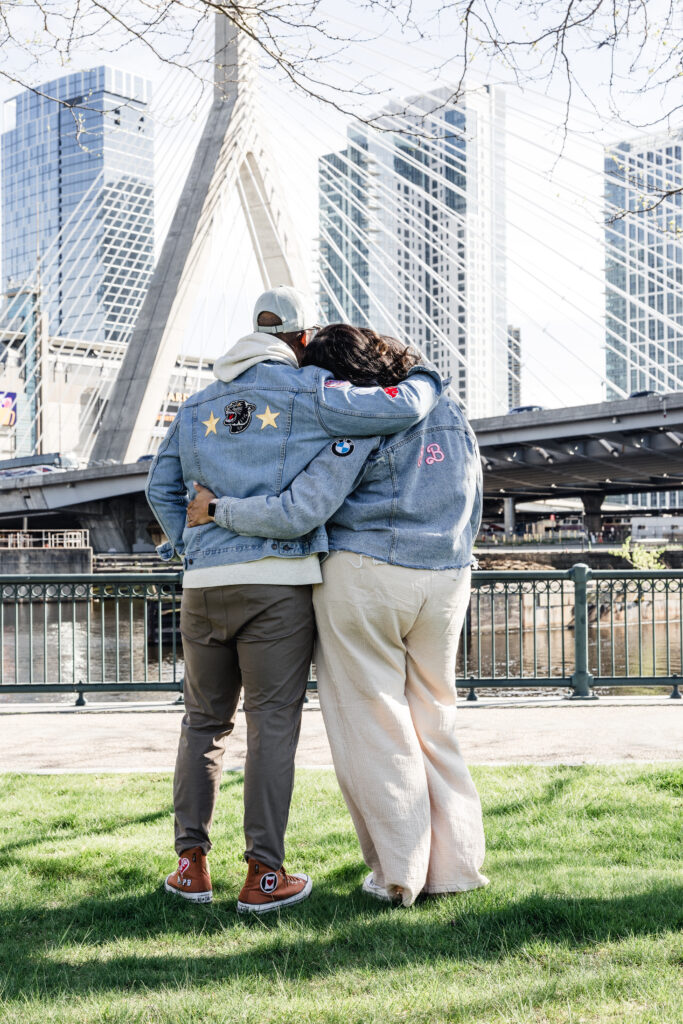 portrait of Palmira and Peterson standing on a grassy area in Boston with skyscrapers and the Zakim Bridge behind them.