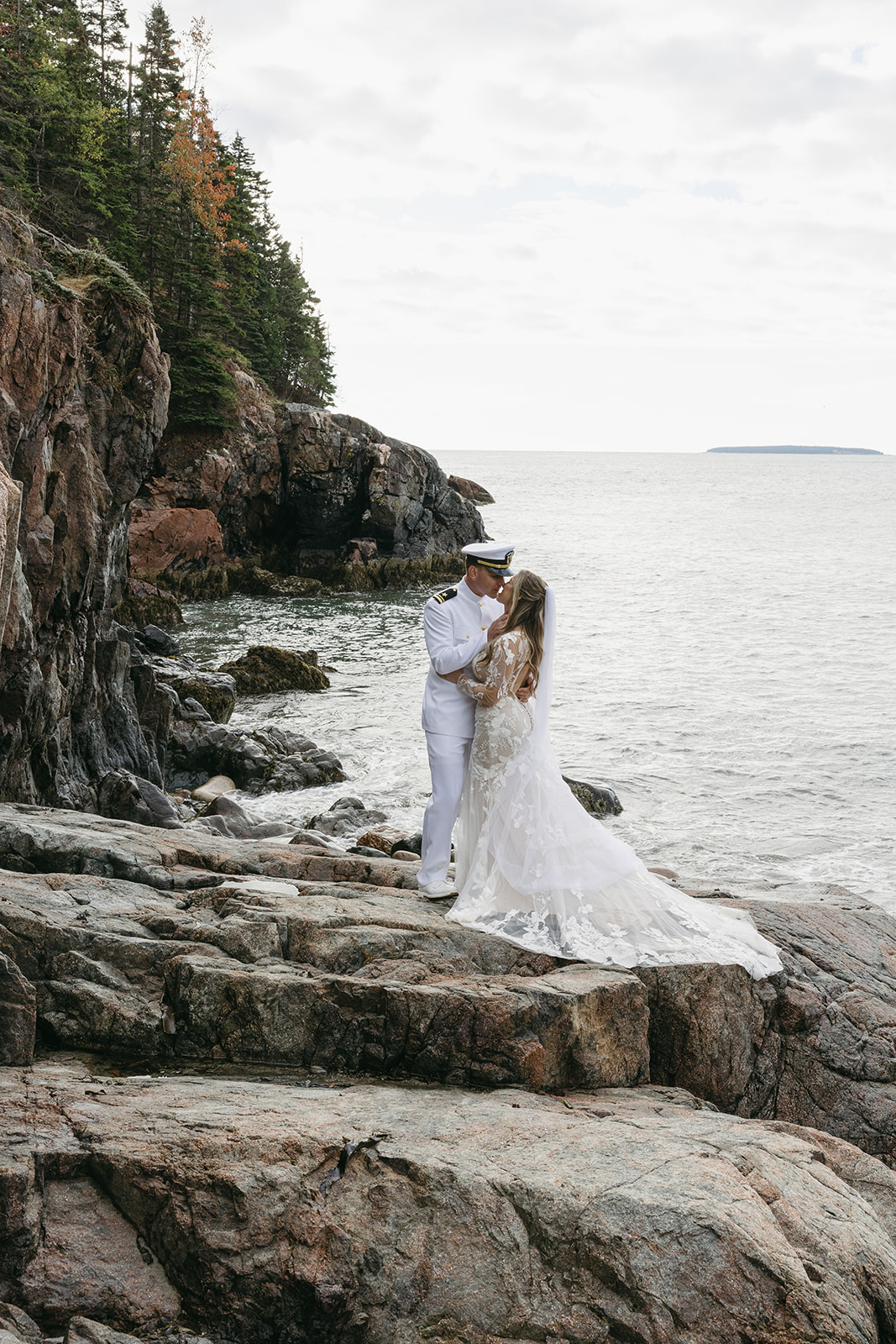 Bride and groom kissing on rocky shoreline in Acadia National Park, with the groom in Navy dress whites and the bride in a floral lace gown.
