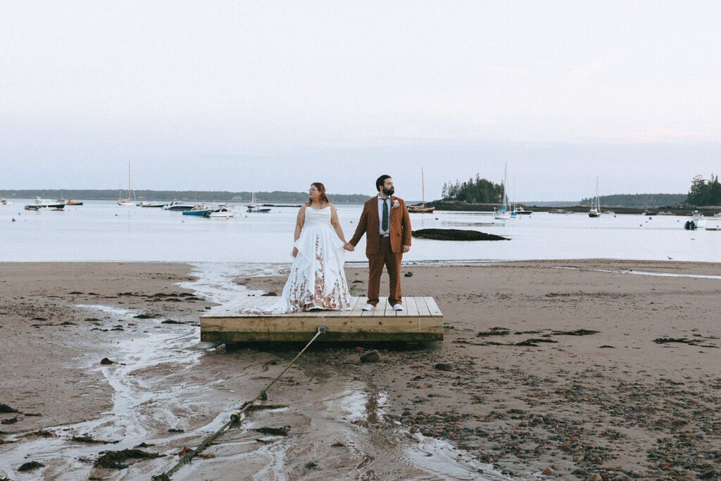 Newlywed couple holding hands on a dock overlooking a Maine harbor filled with sailboats