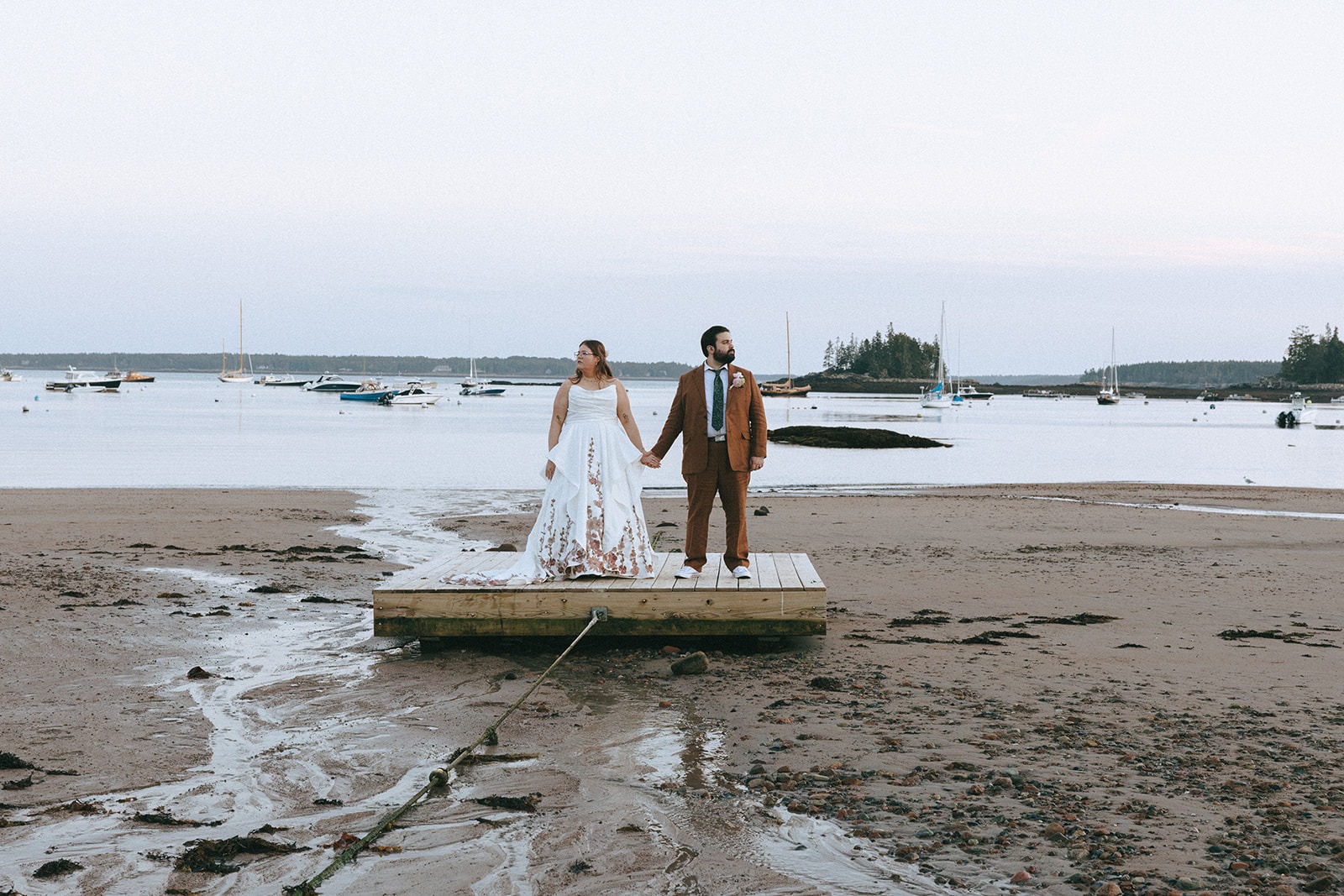 Newlywed couple holding hands on a dock overlooking a Maine harbor filled with sailboats